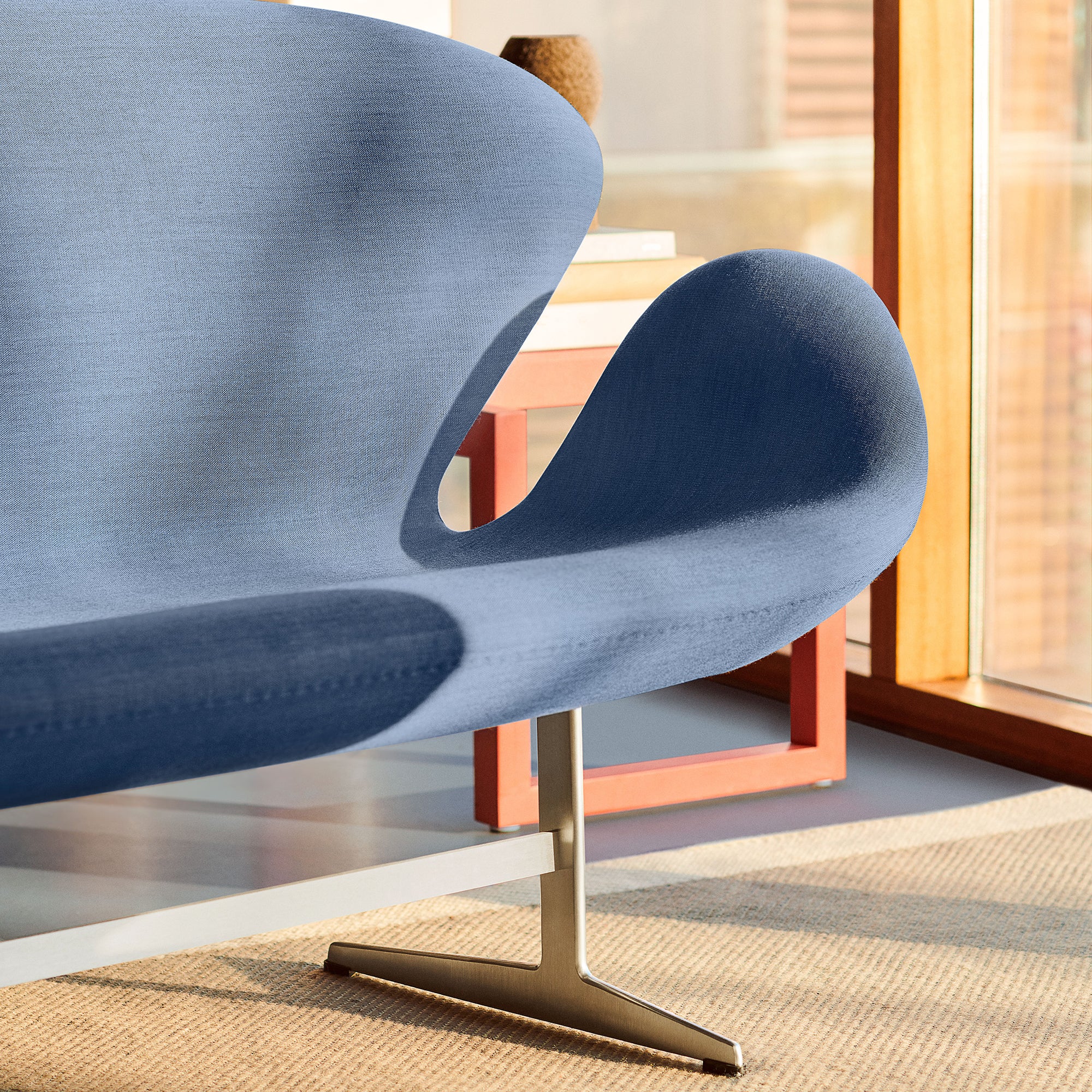 Close-up of a curved blue fabric sofa on a satin metal pedestal base, beside a red-orange side table