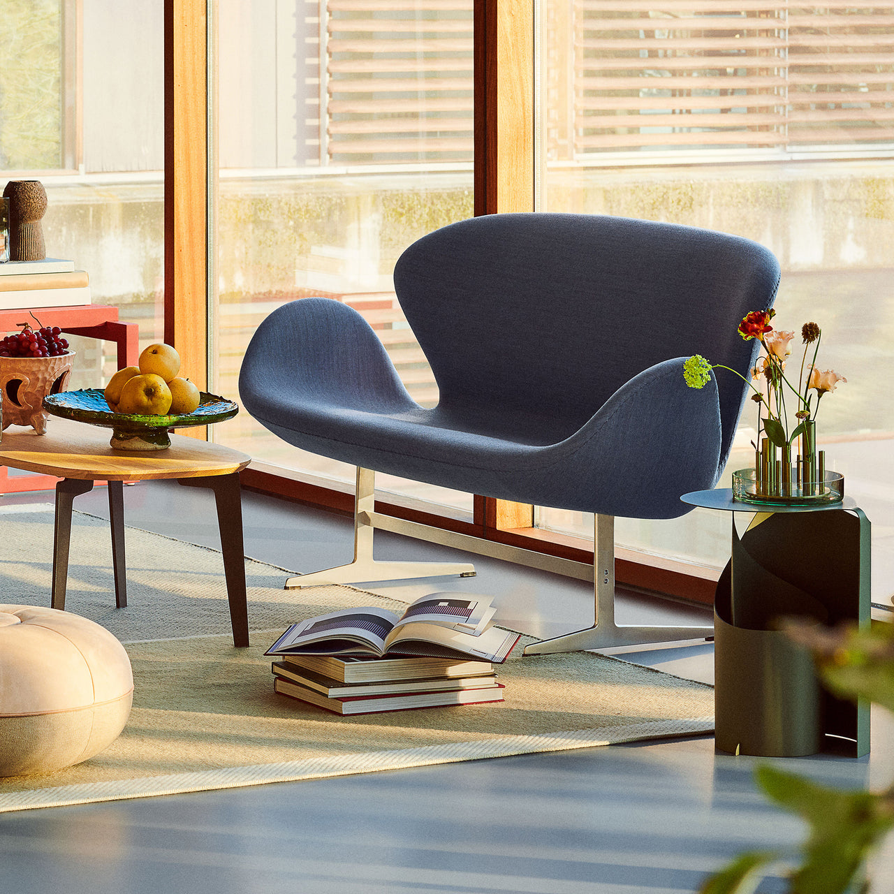 A curved two-seat sofa in blue-grey fabric on a pale metal base, beside a wooden side table by large windows