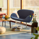 A curved two-seat sofa in blue-grey fabric on a pale metal base, beside a wooden side table by large windows