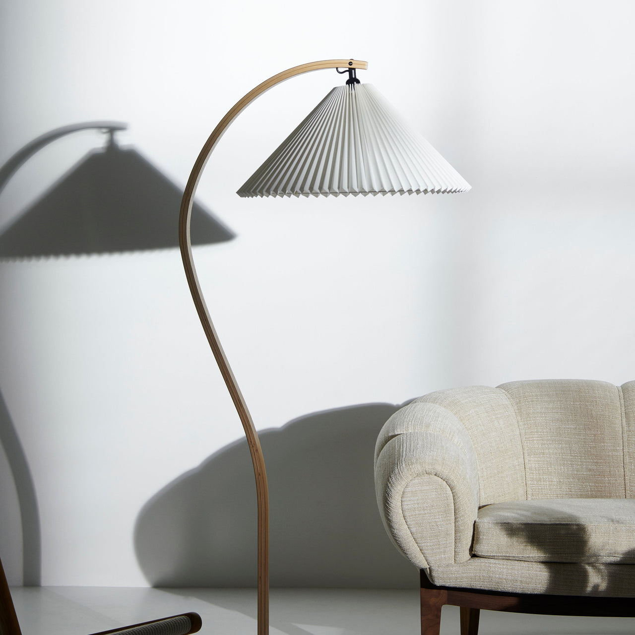 A floor lamp with a curved light wood arm and pleated white shade, casting shadows beside a cream sofa