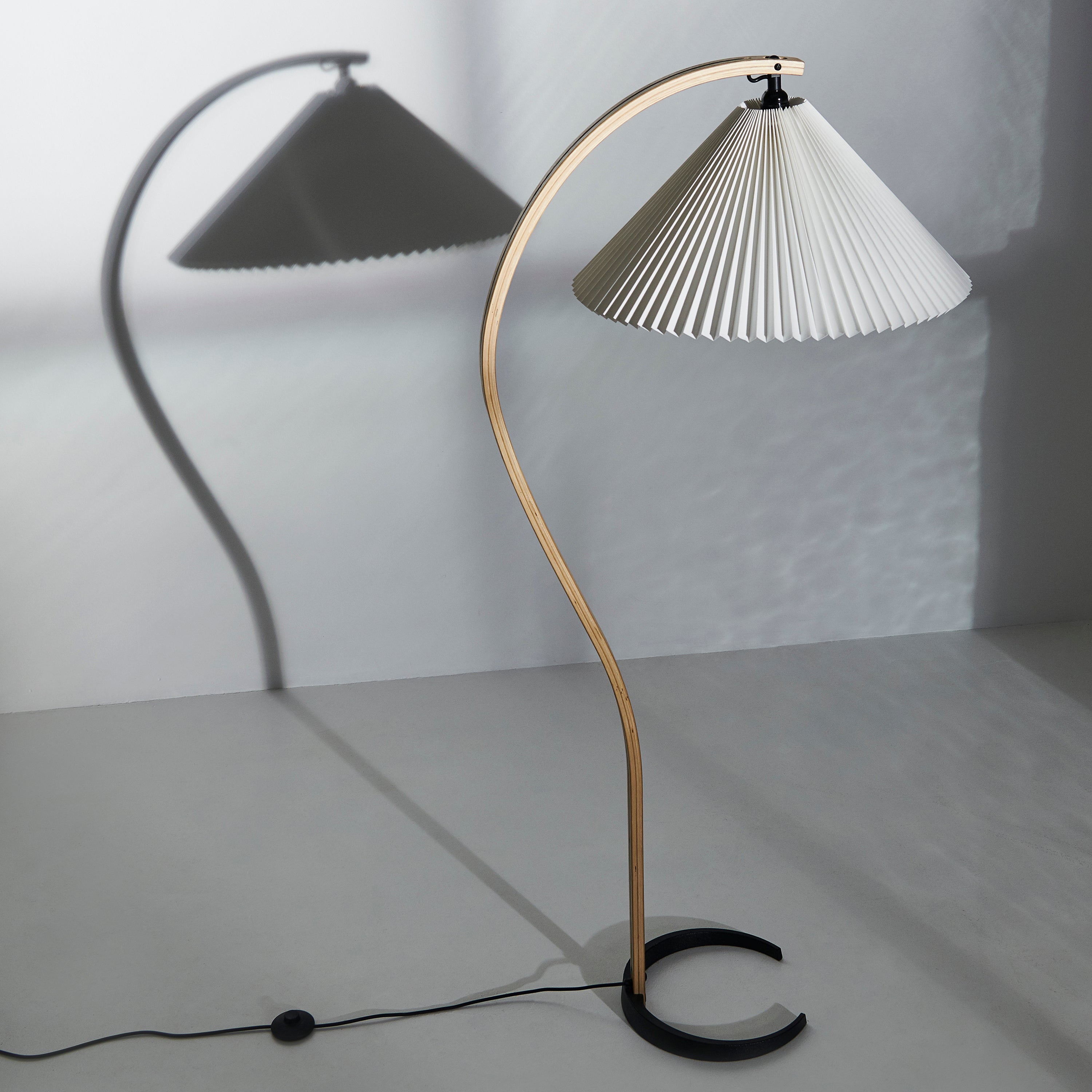 A floor lamp with a curved light wood arm, pleated white shade and black crescent base, casting a shadow on the wall