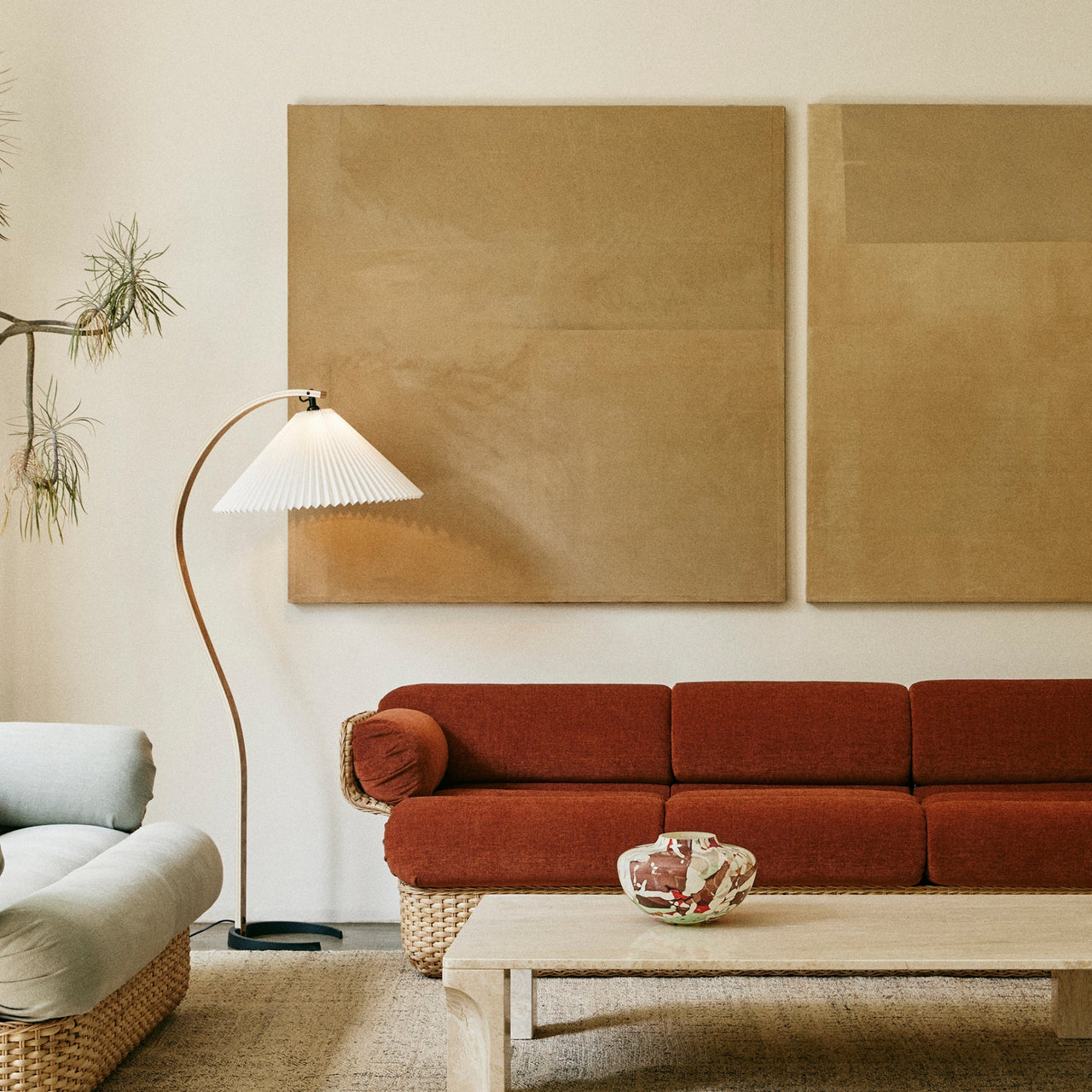 A curved wooden floor lamp with a white pleated shade beside a rust-red sofa in a living room
