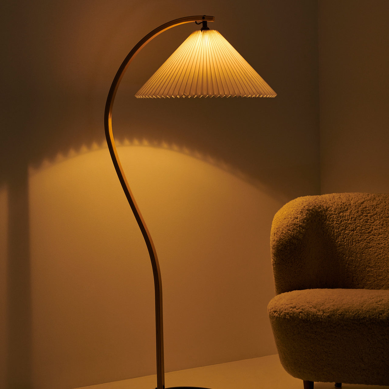 A glowing floor lamp with a curved wooden stem and pleated shade beside a fluffy tan armchair