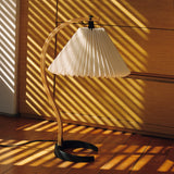 A table lamp with a pleated white shade and curved wooden arm on a wood floor in striped sunlight