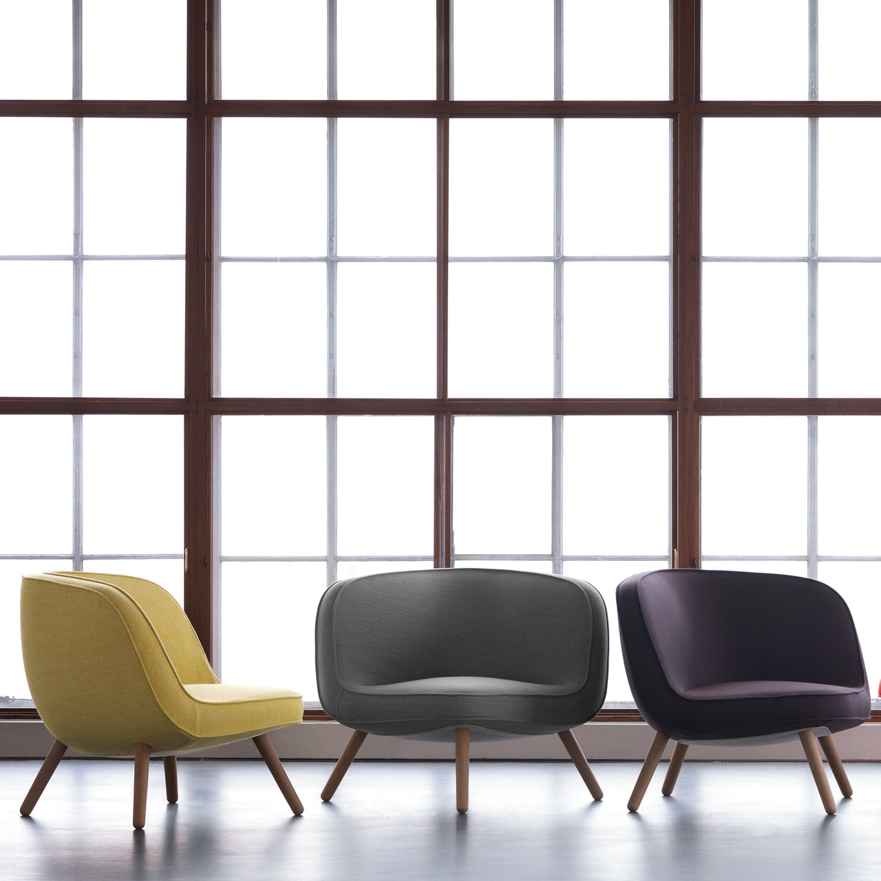 Three low upholstered lounge chairs in yellow, grey and dark plum on slim wooden legs by a large window