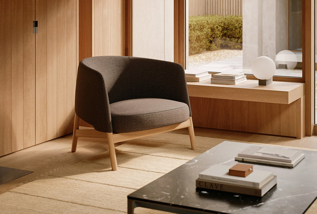 A modern upholstered armchair with a dark gray fabric and light wood legs, placed in a minimalist, warm-toned interior with wood paneling, a low wooden bench, and a marble coffee table.
