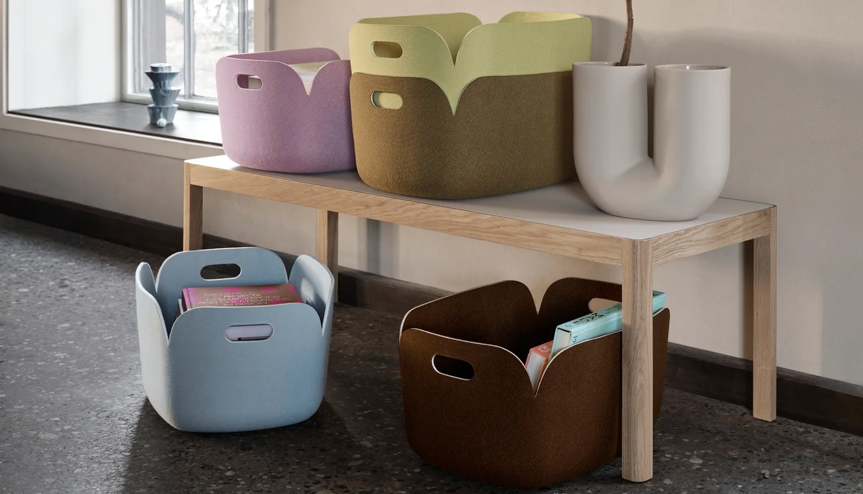 Four fabric storage baskets in pastel and earthy tones arranged on and around a light wood bench in a real interior room with a window, floor, and wall.
