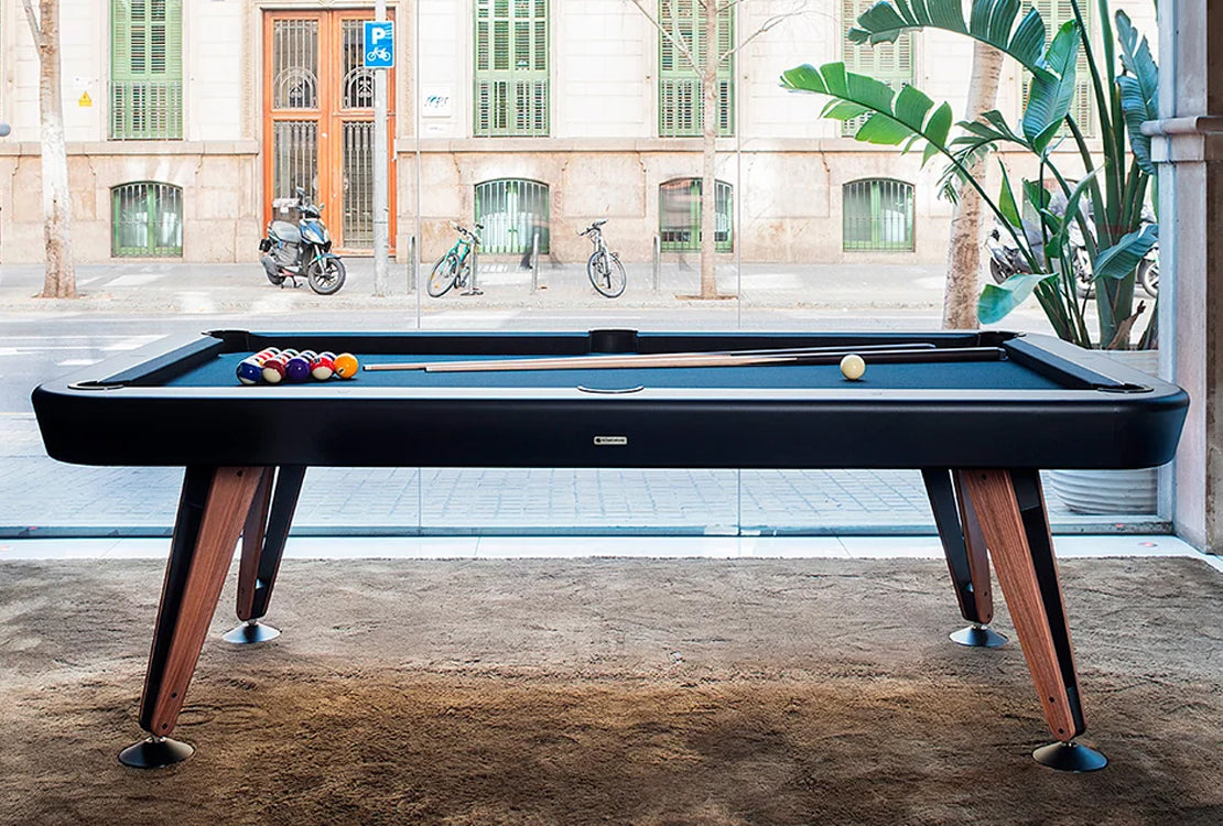 A modern pool table with a black felt surface and dark wood legs, positioned in a bright room with a large window overlooking a city street with bicycles and buildings.