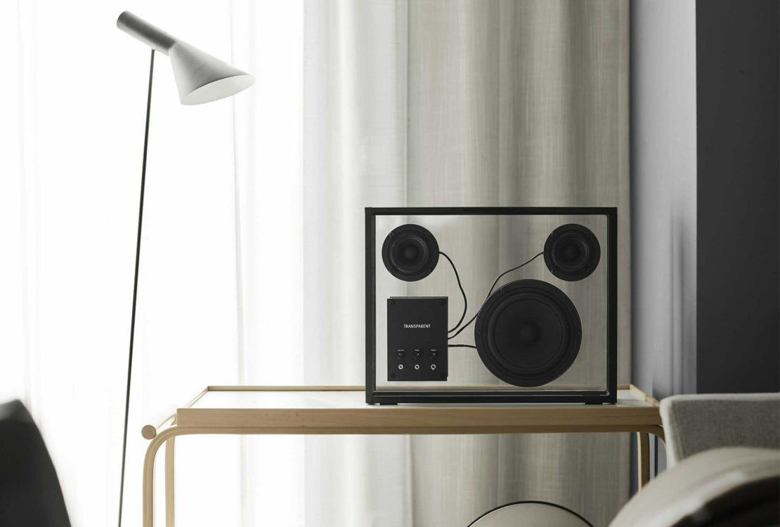 A rectangular black and silver speaker system with three circular drivers sits on a light wood table in a modern room with sheer curtains, a floor lamp, and a chair partially visible.