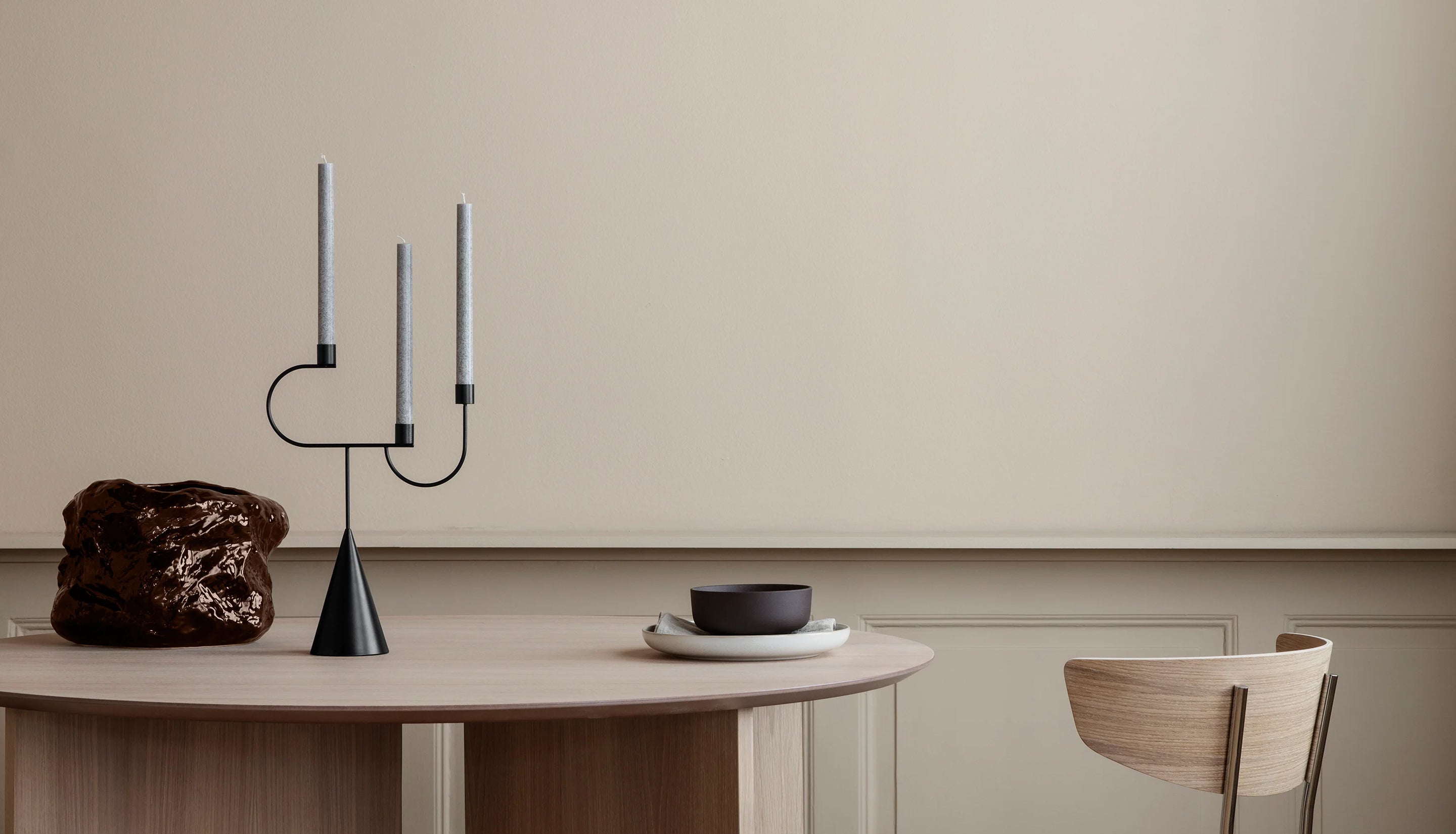 A minimalist dining table setup in a warm neutral room, featuring a black metal three-arm candelabra with white taper candles, a dark brown textured sculpture, a small black bowl on a white plate, and a light wood chair against a beige wall.
