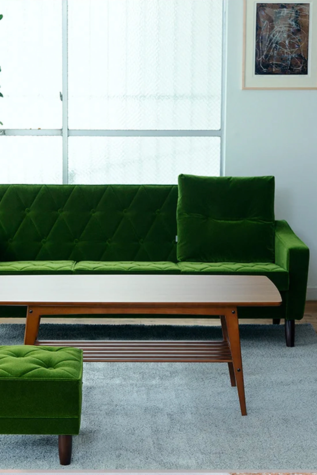 A green velvet tufted sofa with matching cushions in a bright living room, paired with a wooden coffee table and a matching green ottoman in the foreground.