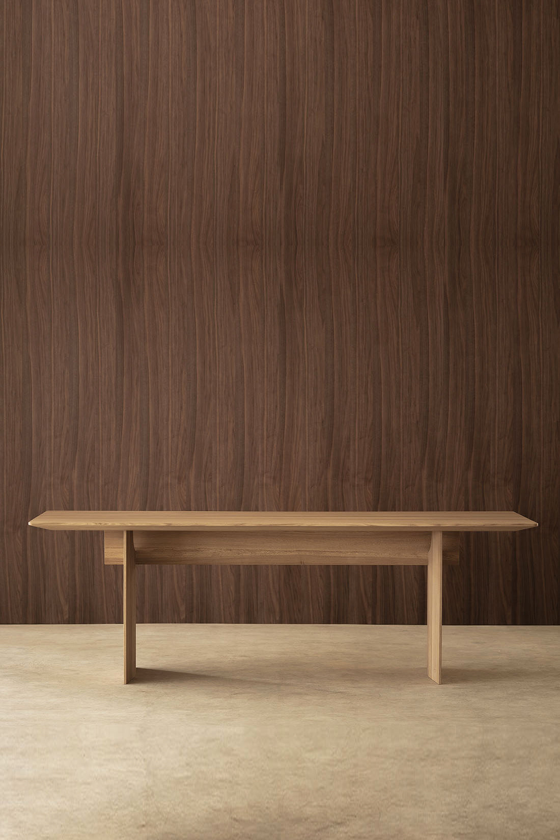 A minimalist light oak wooden console table with a long rectangular top and four tapered legs, set against a dark brown wood-paneled wall and a light concrete floor.