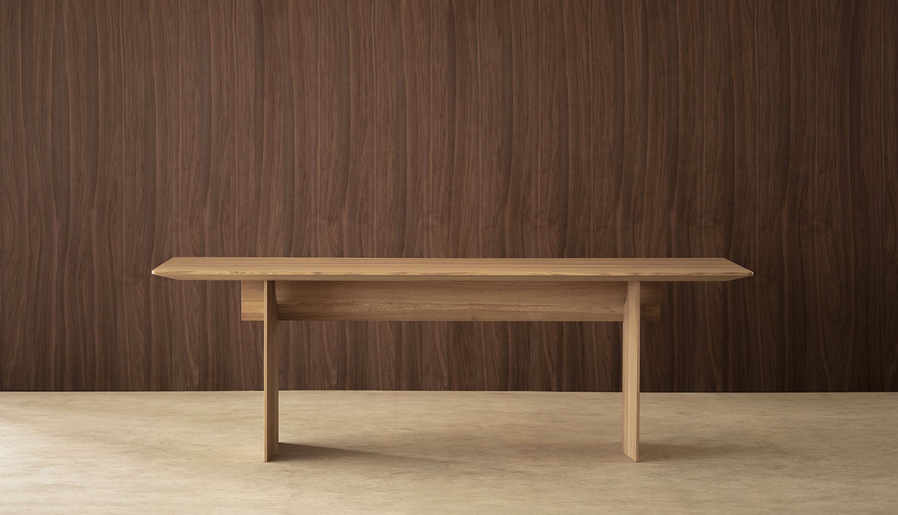 A minimalist light oak wooden dining table with a rectangular top and four tapered legs, set against a dark brown wood-paneled wall and a light concrete floor.