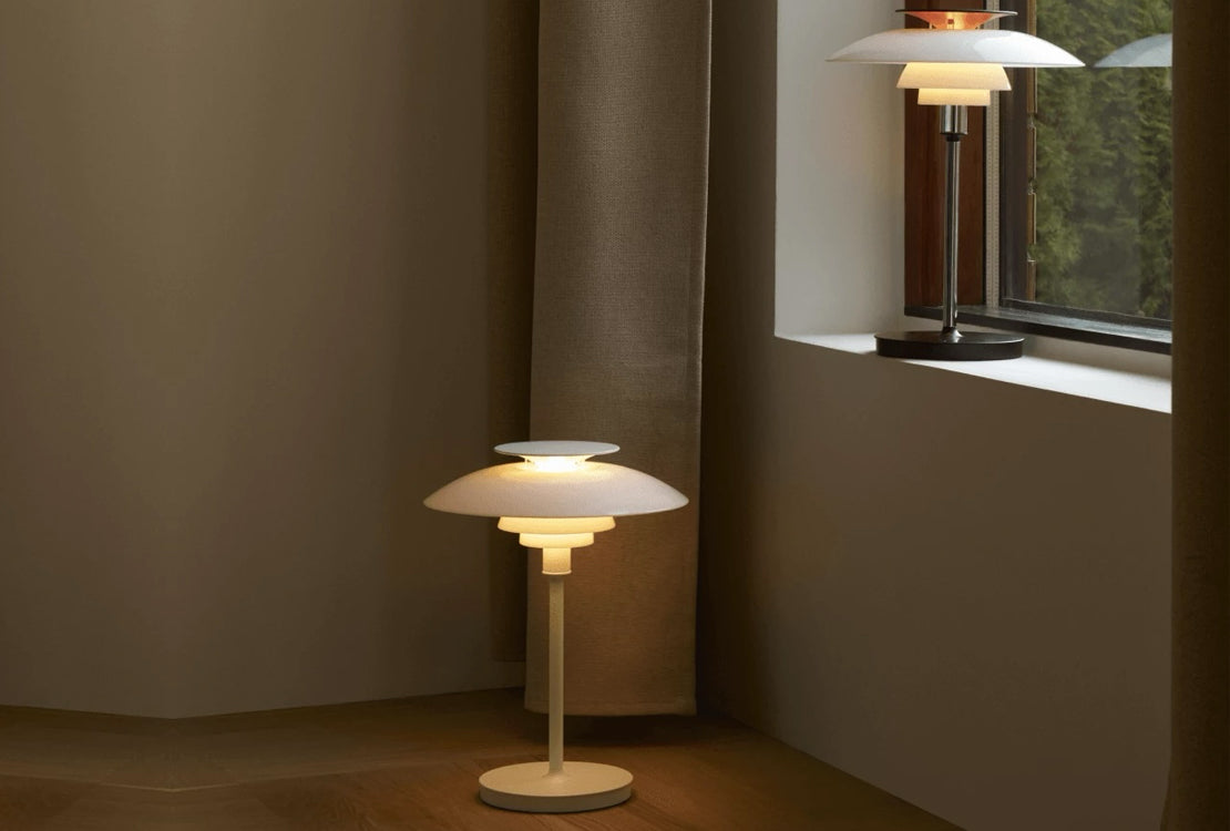 A modern mushroom-shaped table lamp with a warm white glowing shade and stem, placed on a wooden floor in a minimalist room corner. A second, unlit version of the same lamp is visible on a windowsill in the background.