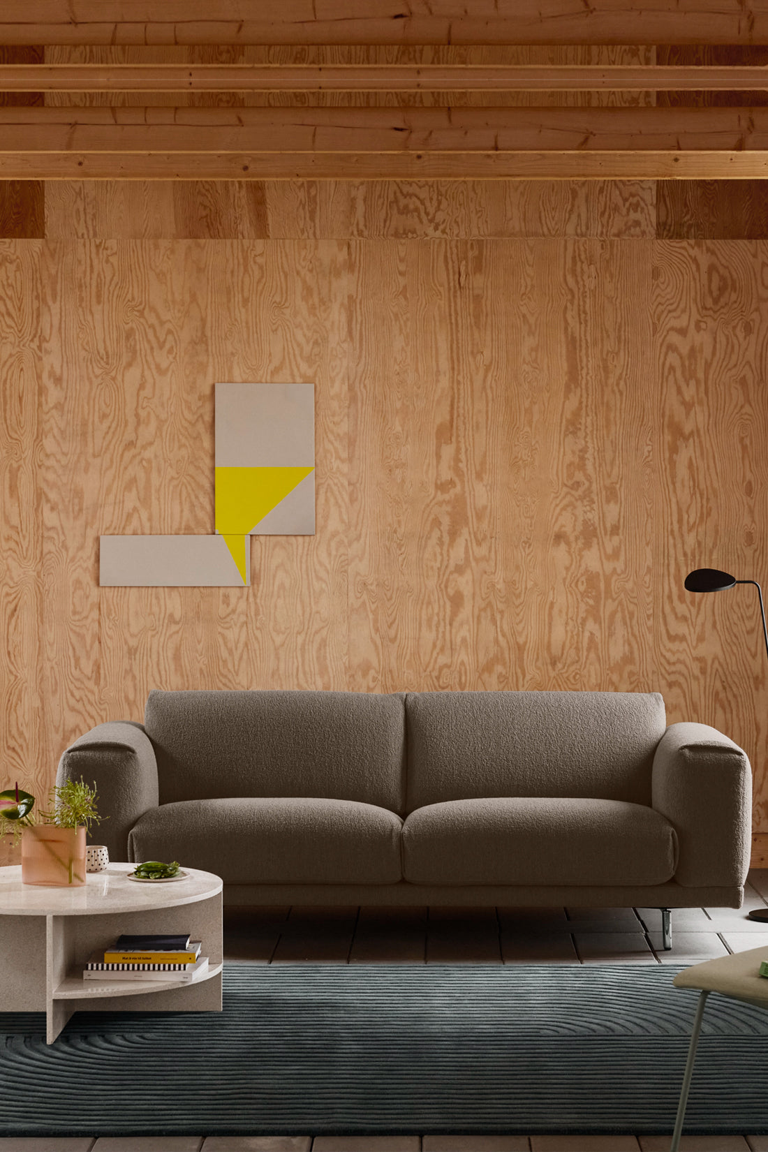 A modern gray fabric sofa set against a light wood plywood wall in a minimalist living room, with a white coffee table, a dark blue textured rug, and abstract geometric wall art.