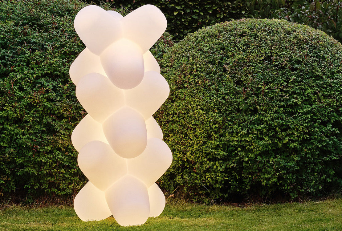 A modern outdoor LED light sculpture shaped like a stacked column of rounded, bubble-like forms, glowing warm white against a background of green garden hedges and grass.