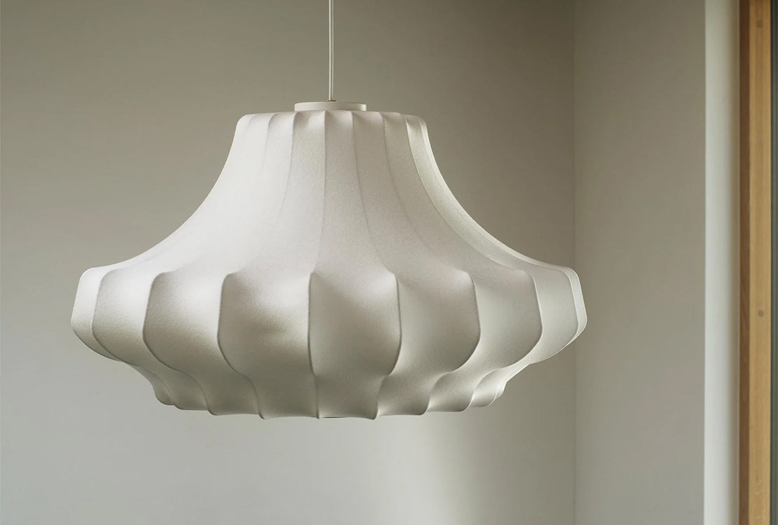 A white pendant light with a sculptural, fluted shade hanging from a thin cord in a minimalist room corner.