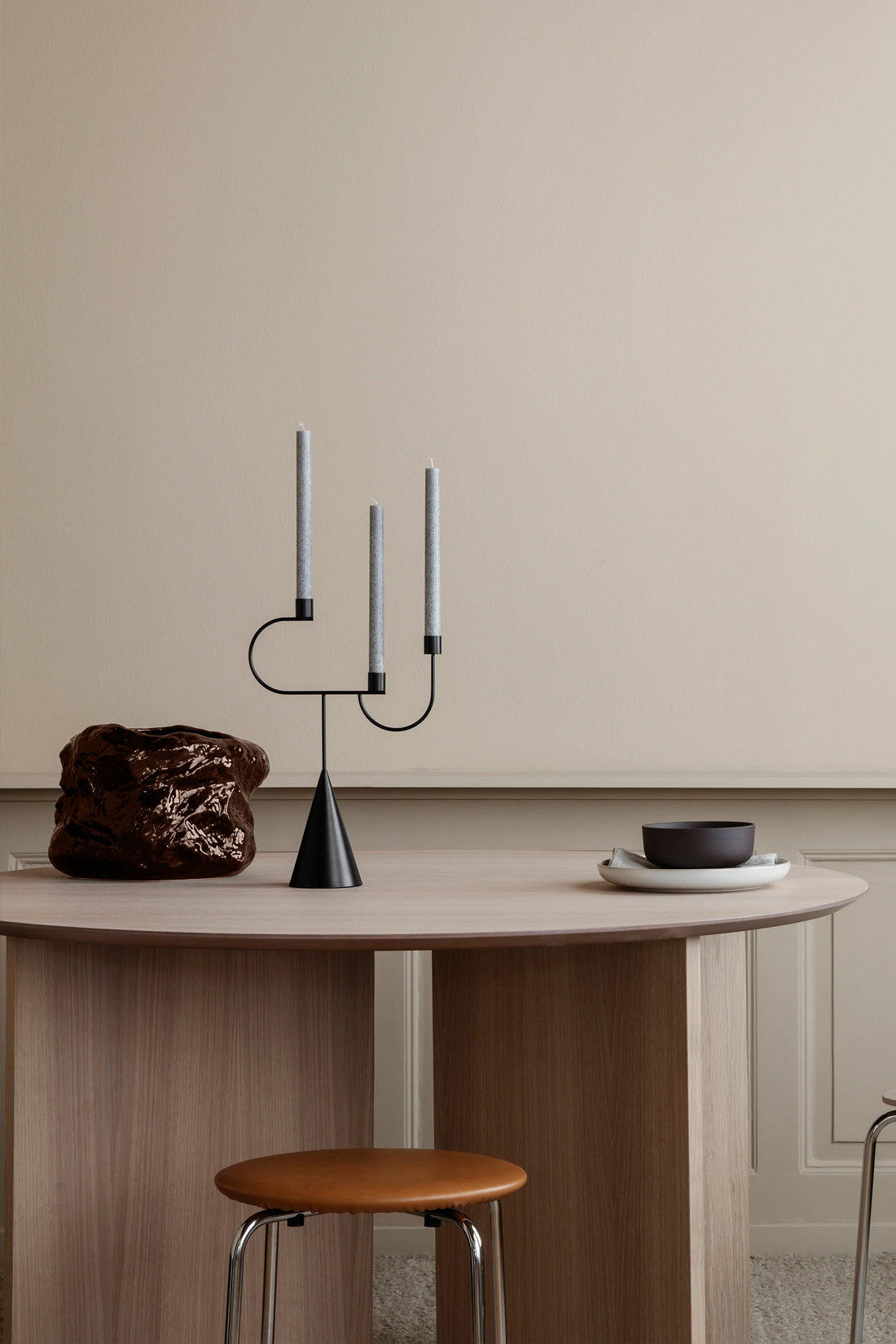 A modern black metal three-arm candelabra with slim white taper candles sits on a light wood dining table in a minimalist room setting, alongside a brown textured vase and a black bowl on a saucer.