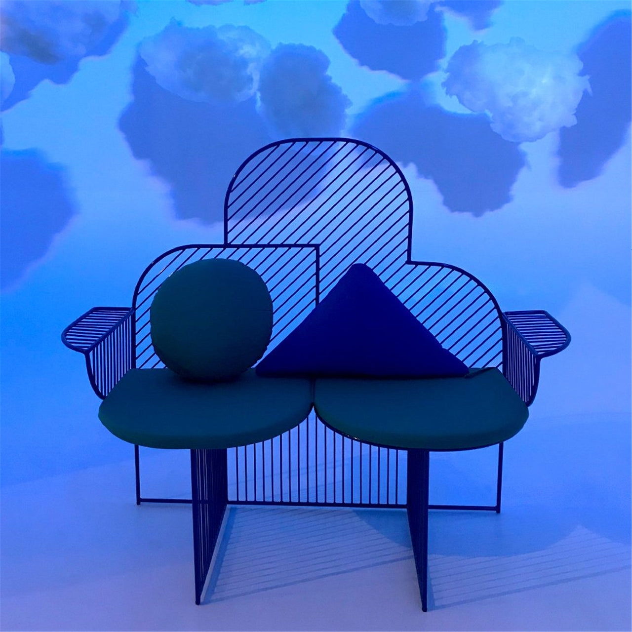 Electric blue metal Cloud Bench with rounded backrest and abstract green and blue cushions against blue cloud backdrop.