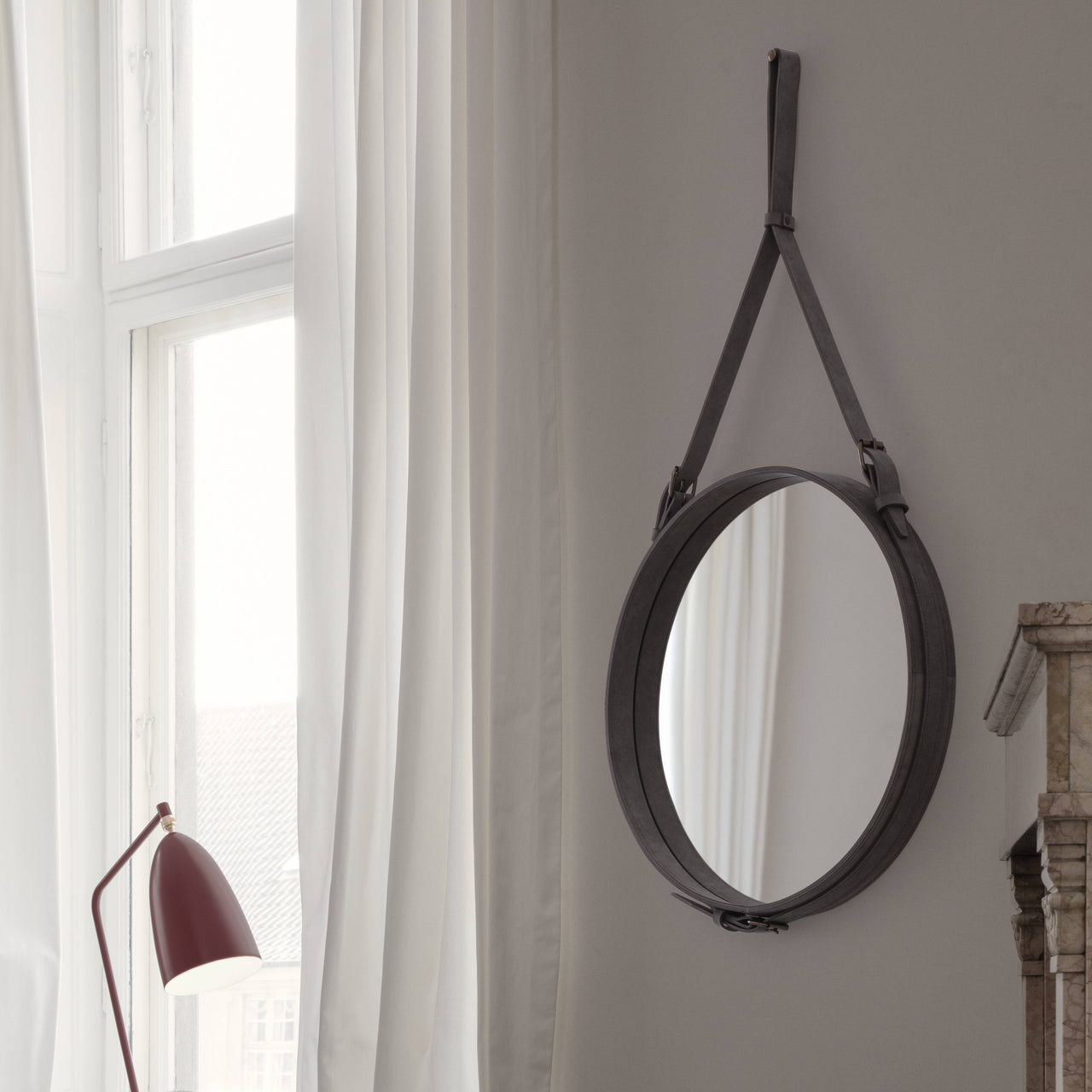 A round mirror with a dark leather-wrapped frame hangs from a strap on a pale wall near a window