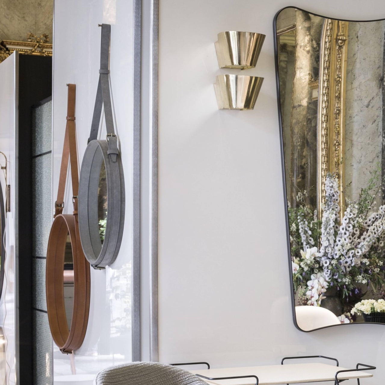 Two round mirrors with leather strap hangers beside a tall dark-framed mirror and brass wall lights