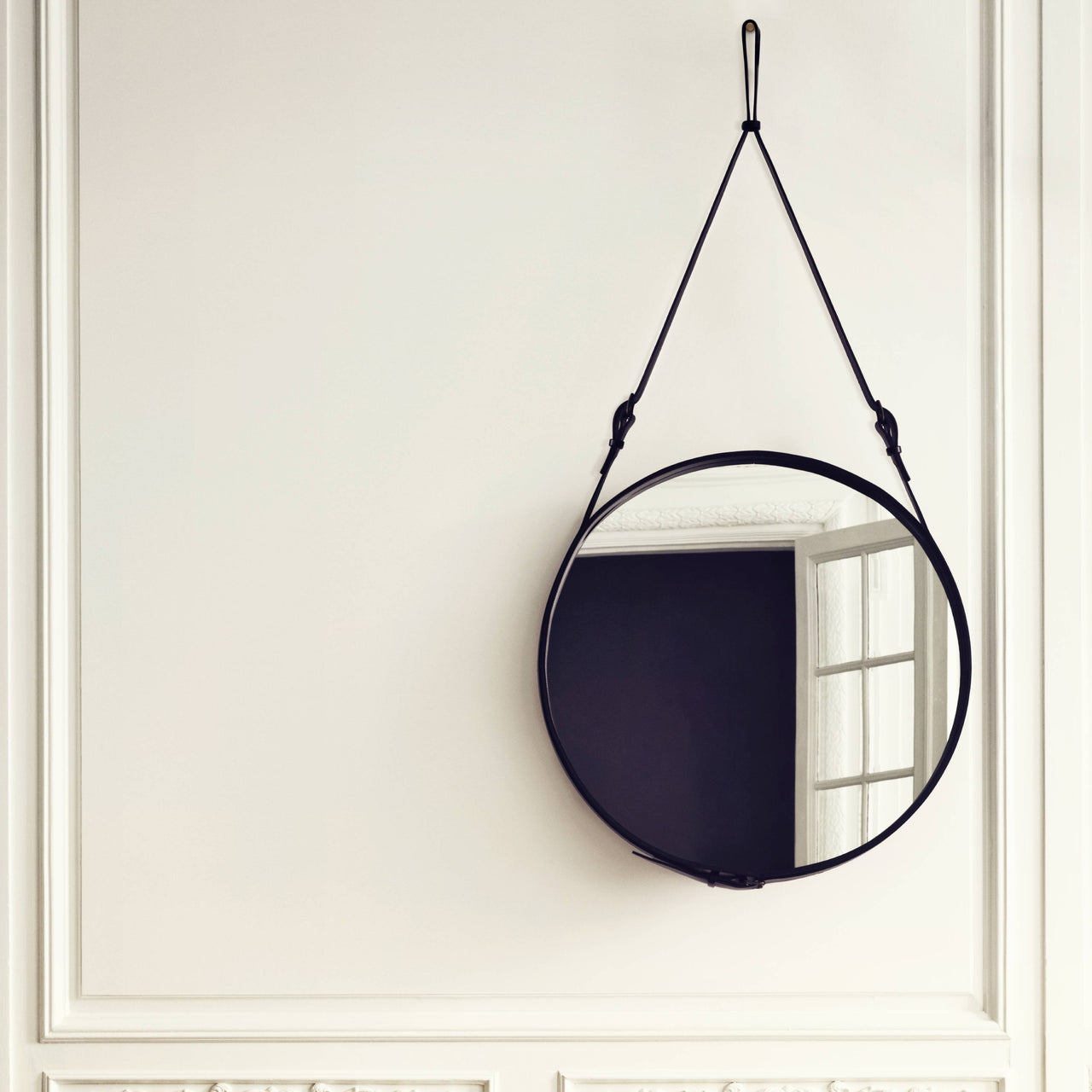 A round mirror in a black leather frame hangs from a strap on a white paneled wall