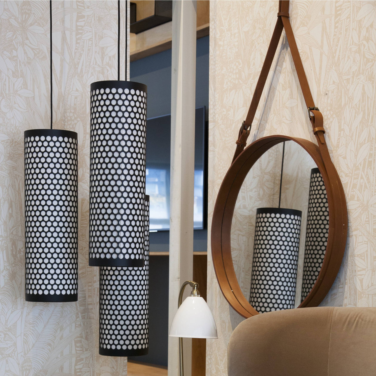 A round mirror with a tan leather frame and strap beside three black perforated cylinder pendant lamps