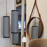 A round mirror with a tan leather frame and strap beside three black perforated cylinder pendant lamps