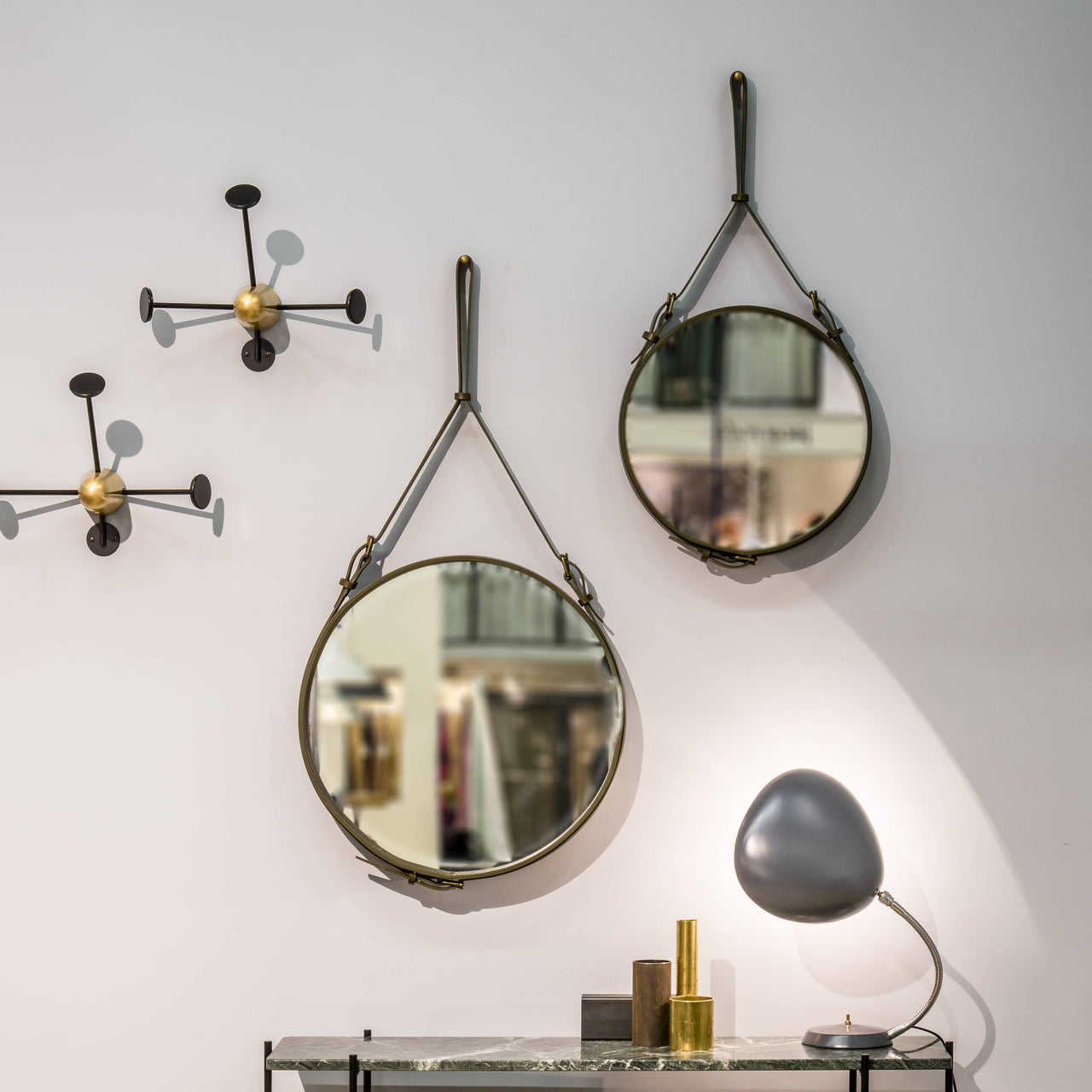 Two round mirrors hung on leather straps on a pale wall, beside black and brass coat hooks