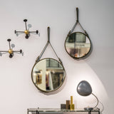 Two round mirrors hung on leather straps on a pale wall, beside black and brass coat hooks