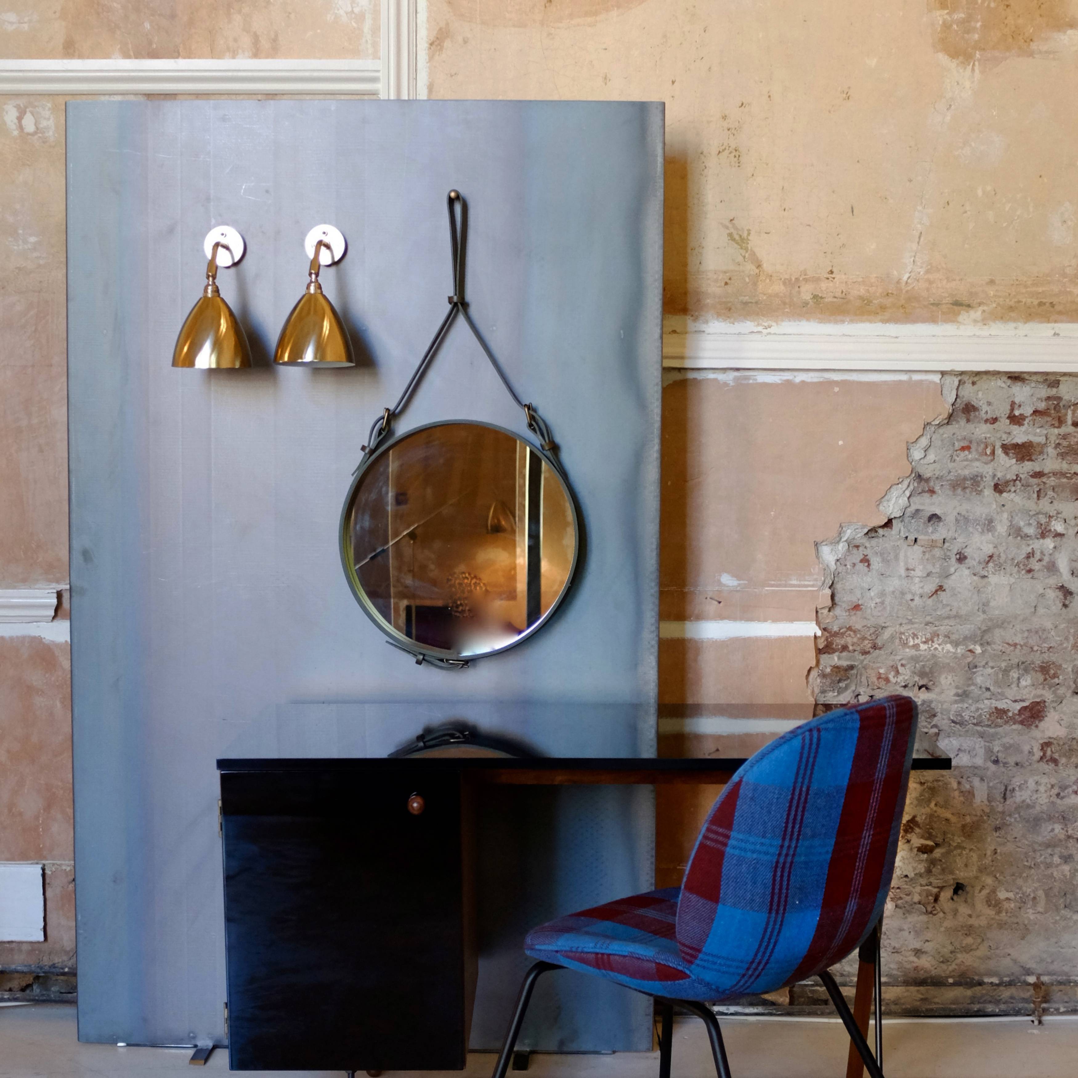 A round mirror hanging from a leather strap beside two brass wall lights, above a dark desk and plaid chair
