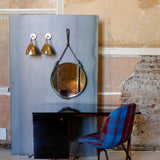 A round mirror hanging from a leather strap beside two brass wall lights, above a dark desk and plaid chair