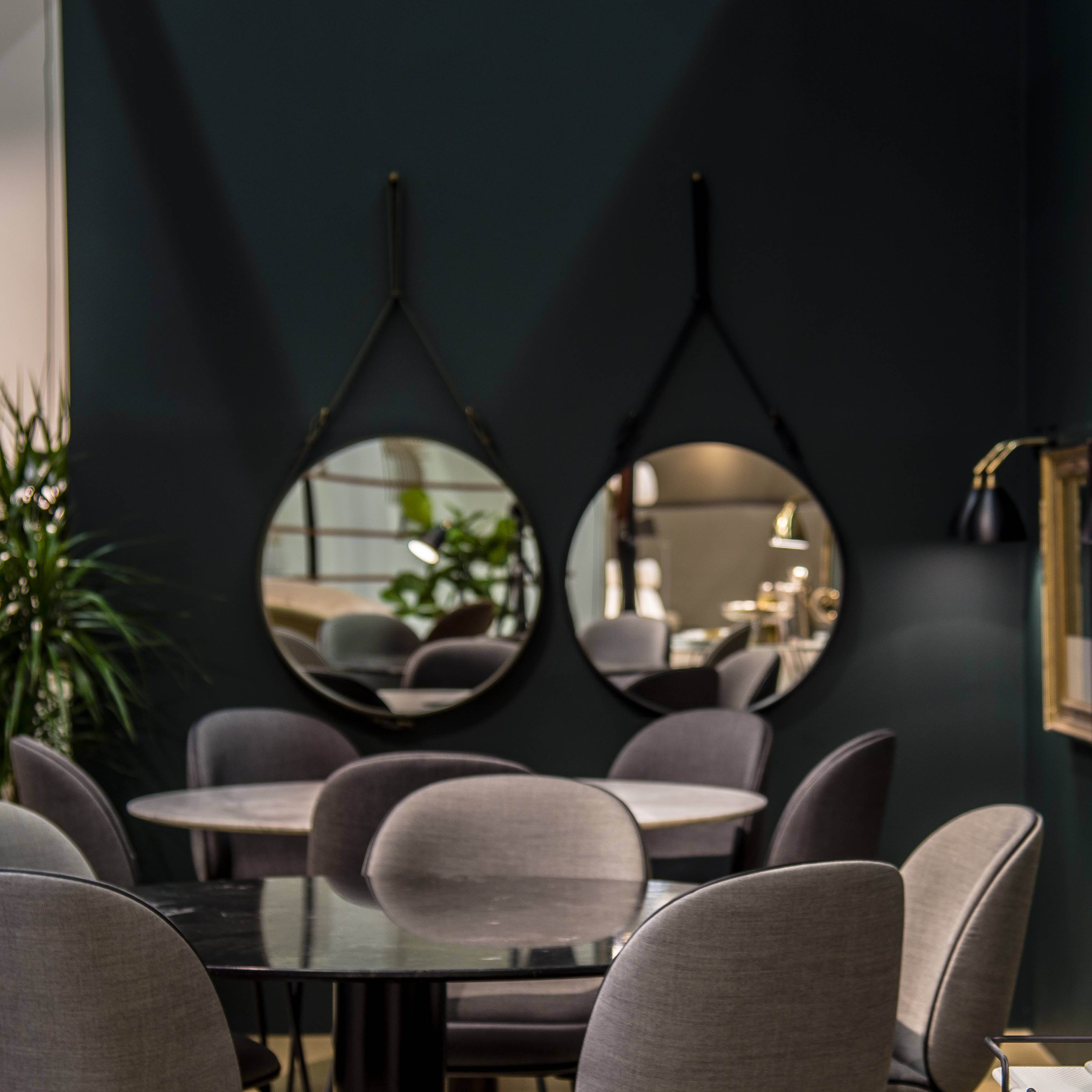 Two round mirrors hang from black leather straps on a dark green wall above grey upholstered chairs and round tables