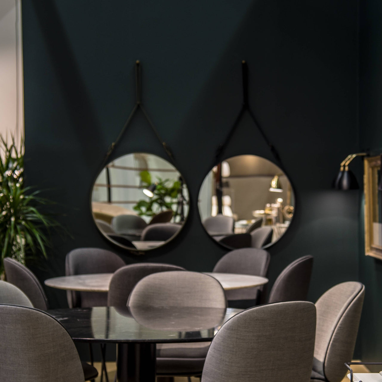 Two round mirrors hang from black leather straps on a dark green wall above grey upholstered chairs and round tables