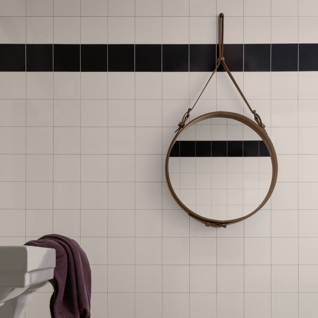 A round mirror in a tan leather frame hangs by a strap on a white tiled wall