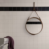 A round mirror in a tan leather frame hangs by a strap on a white tiled wall