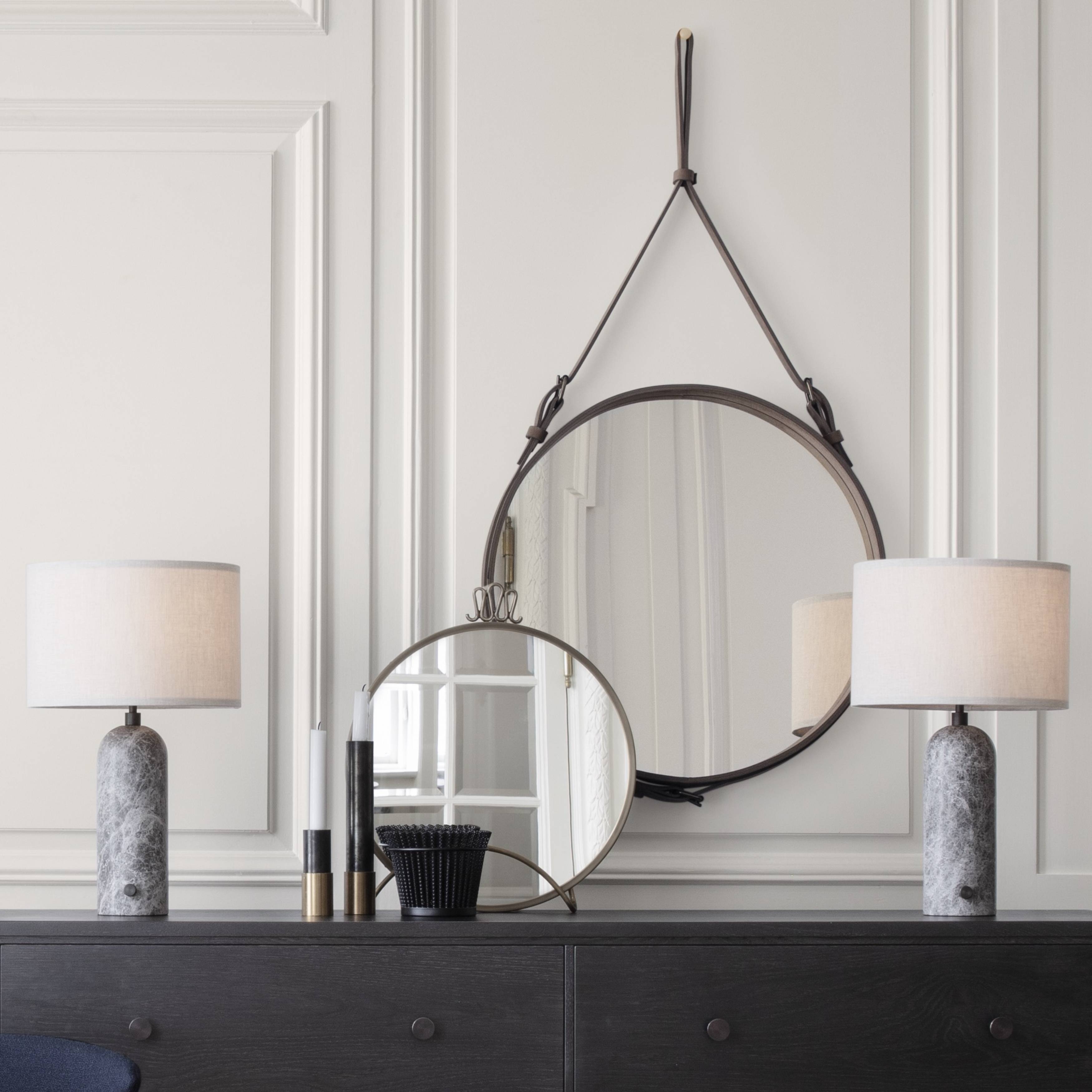 A round mirror hangs from a brown leather strap above a dark dresser with two marble-based lamps