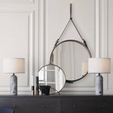 A round mirror hangs from a brown leather strap above a dark dresser with two marble-based lamps
