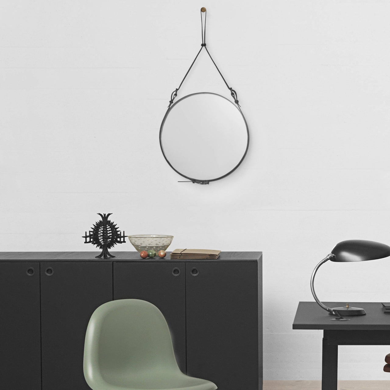 A round mirror with a black strap hangs on a white wall above a dark sideboard and sage green chair