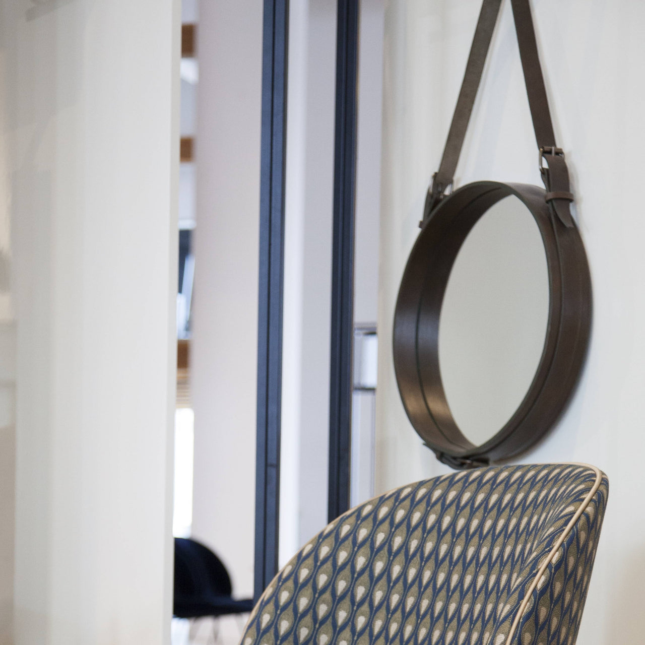 A round mirror with a thick brown leather rim hangs from a strap above a patterned blue chair back