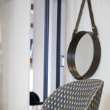 A round mirror with a thick brown leather rim hangs from a strap above a patterned blue chair back