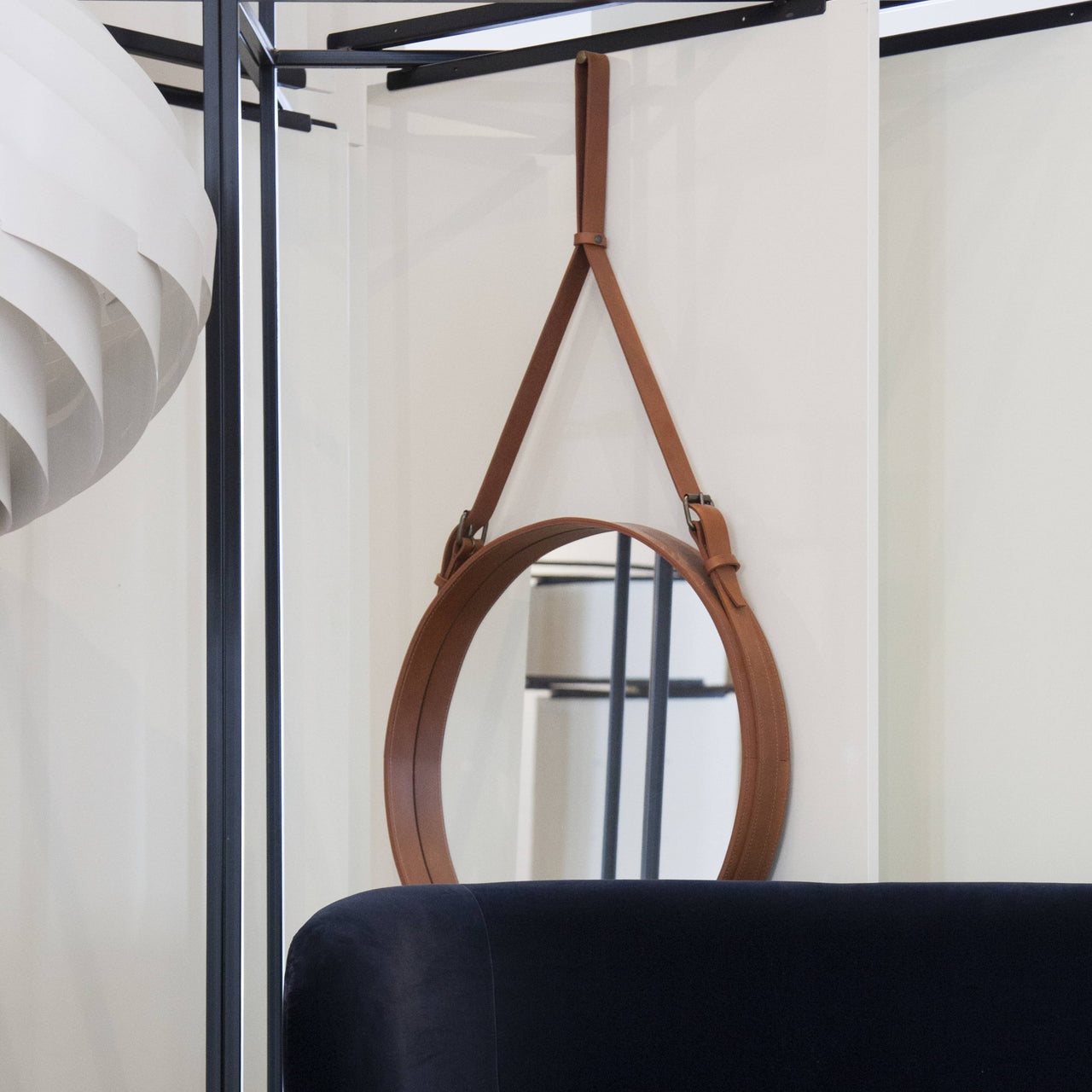 A round mirror with a tan leather frame hangs from a leather strap above a dark blue sofa