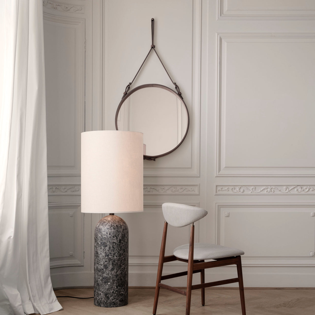 A round mirror hanging from a brown leather strap on a panelled wall, with a marble-based lamp and wooden chair