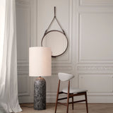 A round mirror hanging from a brown leather strap on a panelled wall, with a marble-based lamp and wooden chair