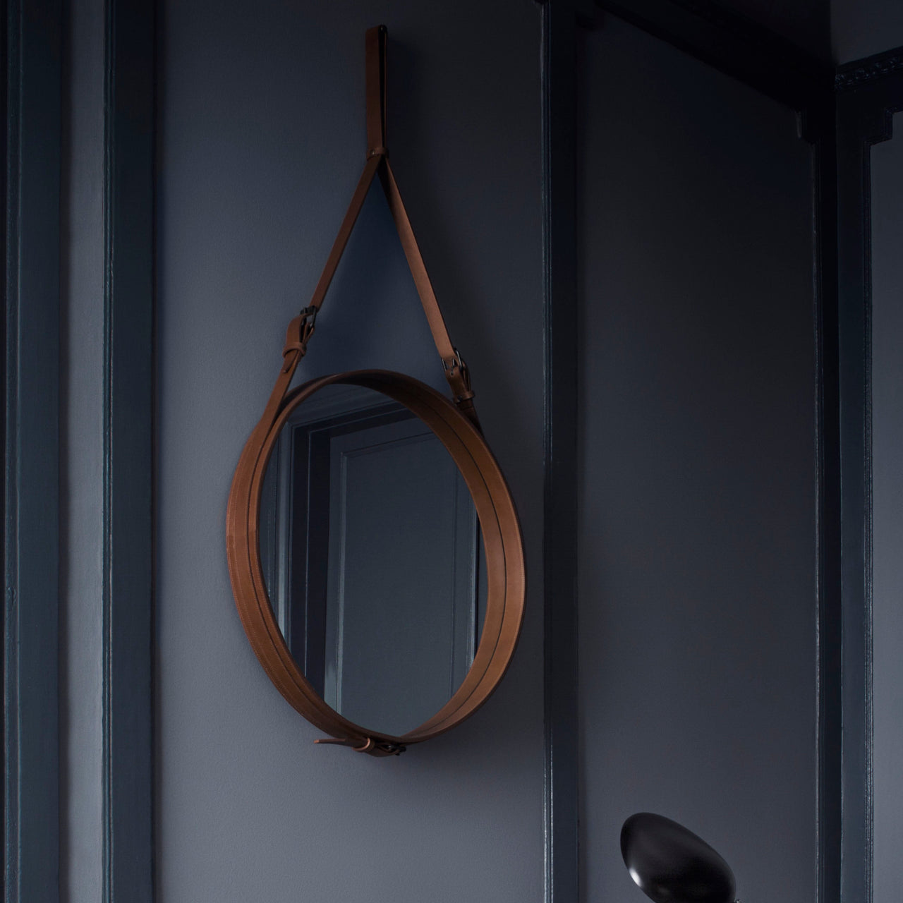 A round mirror in a tan leather frame hangs from a buckled strap on a dark blue-grey wall