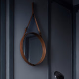 A round mirror in a tan leather frame hangs from a buckled strap on a dark blue-grey wall