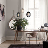 A round mirror with a tan leather strap hangs on a grey wall beside a wooden desk and plant