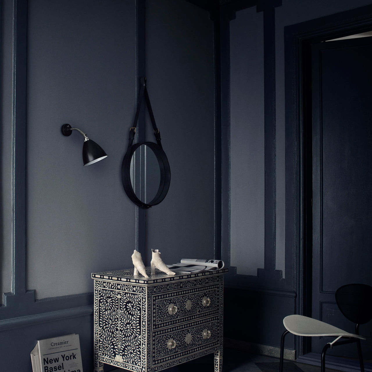 A round mirror with a black leather strap hangs on a navy wall above a patterned chest of drawers
