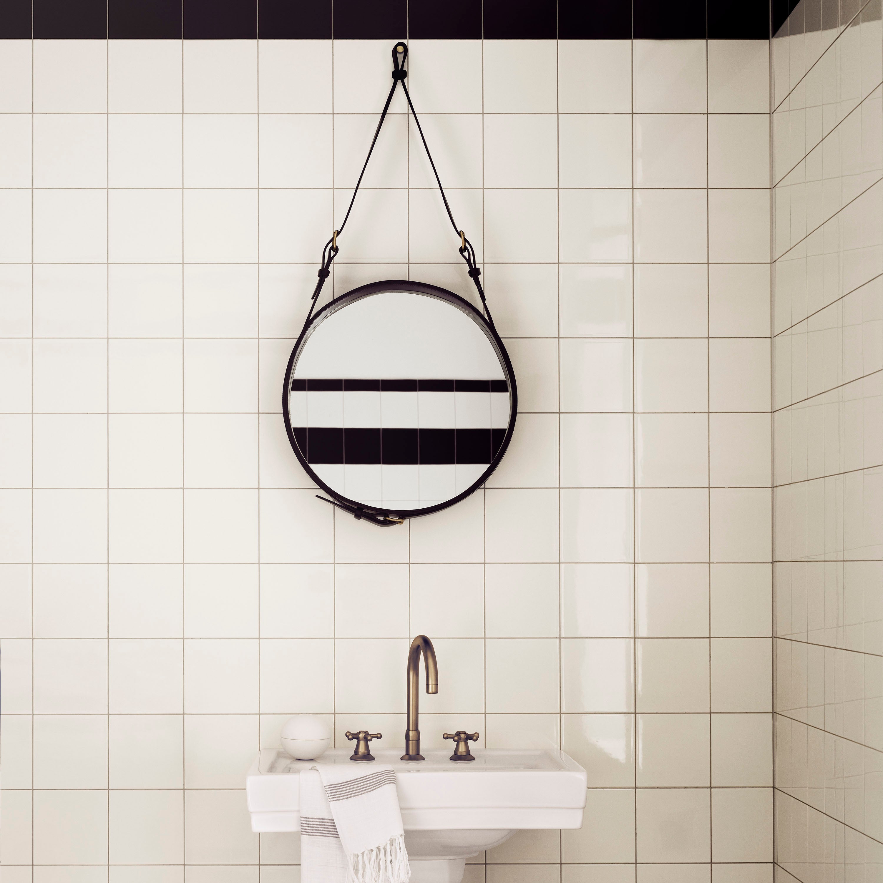 A round mirror with a dark leather strap and edge hangs on a white tiled wall above a pedestal sink