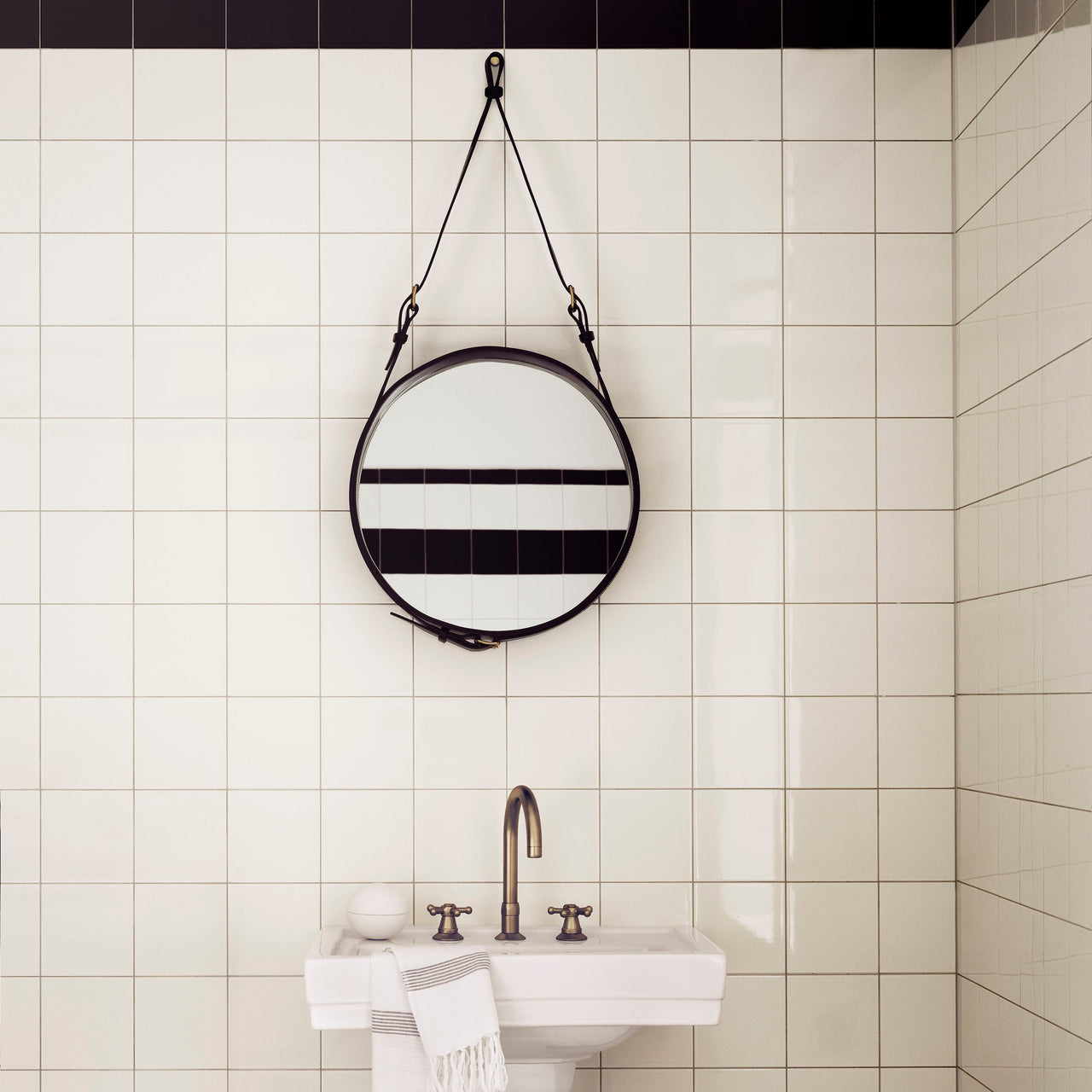 A round mirror with a dark leather strap and edge hangs on a white tiled wall above a pedestal sink